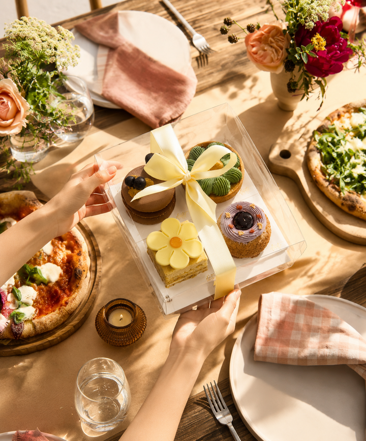 Hands holding an Elevete Patisserie Symphony Dessert Treats gift box with assorted mini desserts including chocolate dome, matcha tart, lemon cake and berry choux, tied with a yellow ribbon, free delivery across Kuala Lumpur and Selangor, halal certified