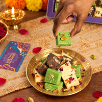 Hand lifting a square of pistachio burfi from a brass plate