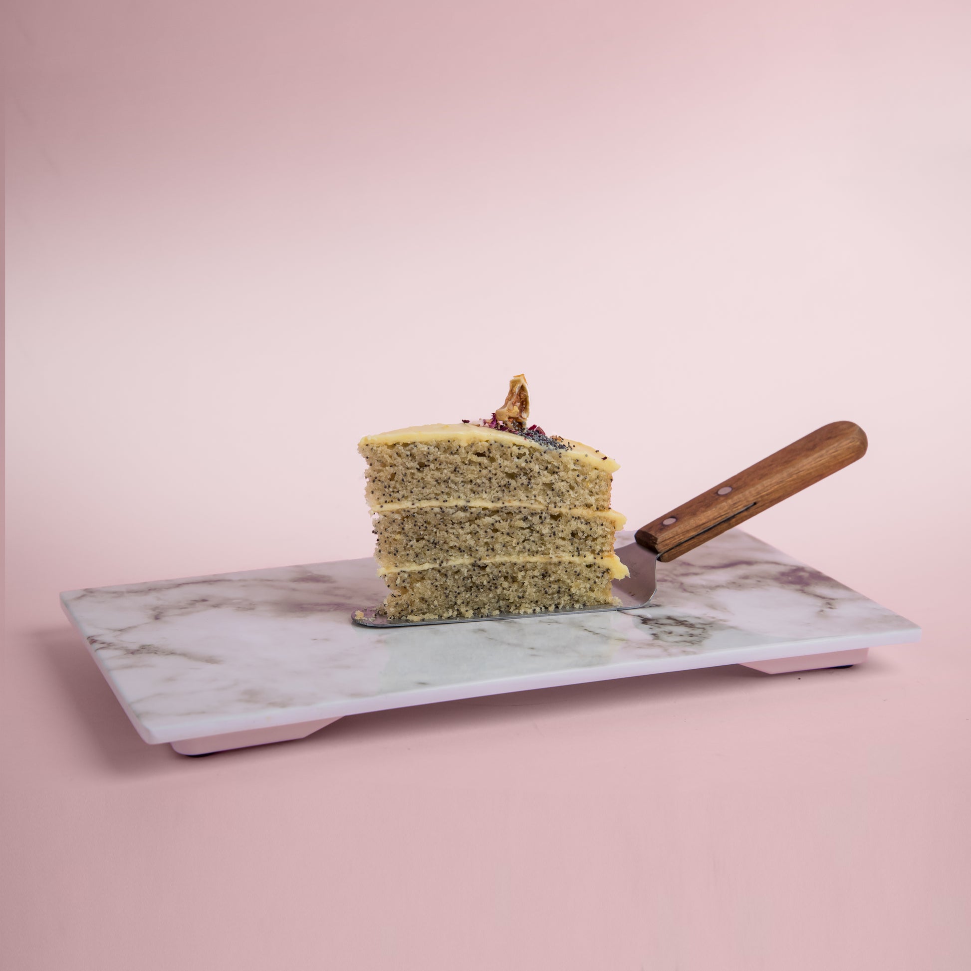 A Serving Slice of Side View of Lemon Poppyseed cake decorated with poppyseed, dehydrated lemon & rose petals on top by Elevete Patisserie